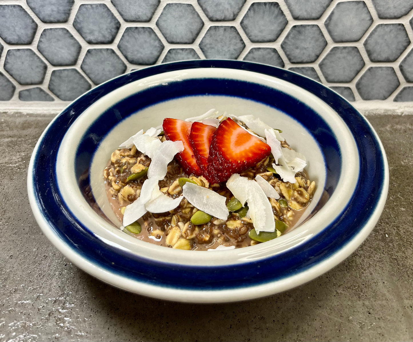 Bowl of Dr. Mak's cacao overnight oats with strawberries and coconut flakes on a gray counter with a tiled background