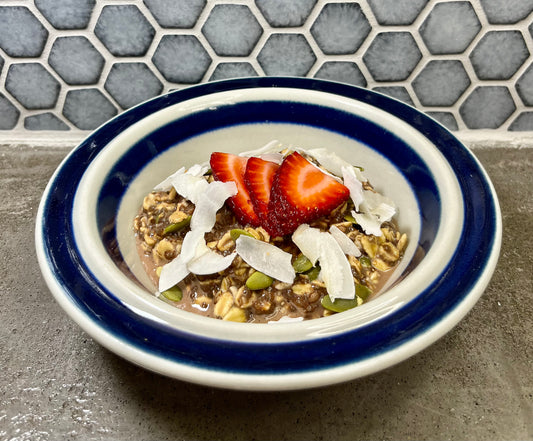 Bowl of Dr. Mak's cacao overnight oats with strawberries and coconut flakes on a gray counter with a tiled background